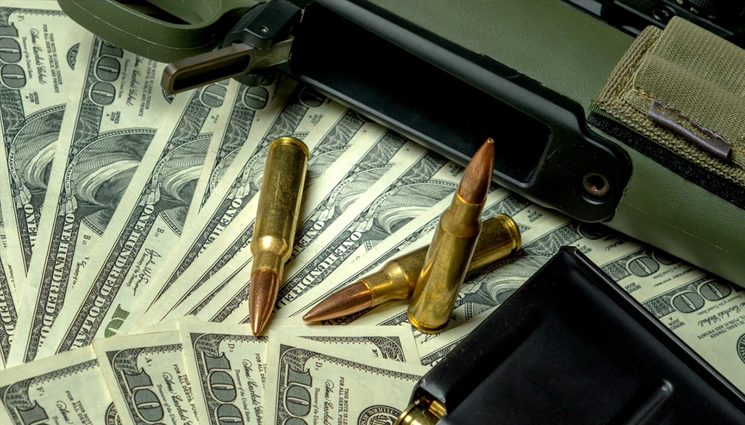 Stack of US dollar bills with rifle ammunition and bullets, representing private military contractor pay and high-threat security work.